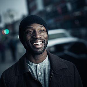 A head and shoulders portrait of a Black man in a black toque, black zib up jacket and grey top who smiles facing the camera. In the background is a blurred out urban setting with pedestrians and traffic.