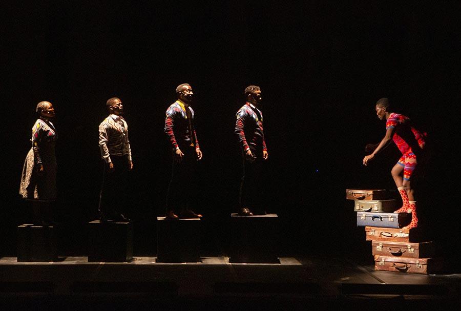 Gregory Maqoma and Thuthuka Sibisi. A Black man in red patterned socks, shorts and t shirt climbs steps made from stacked suitcases. Four other people in colourful clothing face him while standing on boxes in a row. The man on the suitcases is lit brightly, while the others are backlit. Everyone stands out against a black backgound.