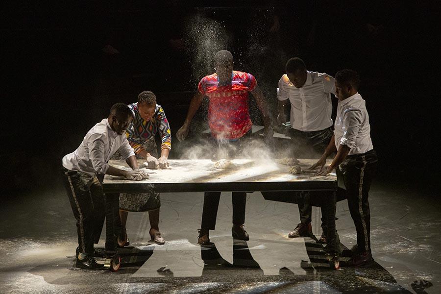 Three Black men dressed in white button up shirts and dark pants, one Black man in a red patterned T-shirt and Black woman in a colourful patterned dress stand around a table covered in flour. There is a lump of dough in the middle and some flour sprays up into the air.