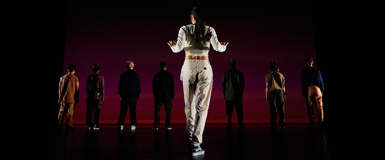 Bboyizm. Light skinned woman with long dark hair, dressed in a long sleeve white crop top, baggy white pants and sneakers stands at the front of a stage with her back to the camera and arms slightly raised. A row of seven people in shadow stand at the back of the stage.