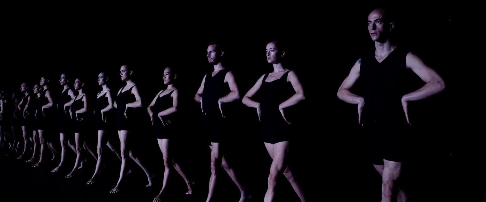 Batsheva Dance Company. Eleven fair skinned dancers in tight black sleeveless tops and shorts stand side by side in a line with their hands on their hips. They are facing slightly to the left of the frame. The dancer on the far right is closest to the camera with the others receding back into the frame to the left.