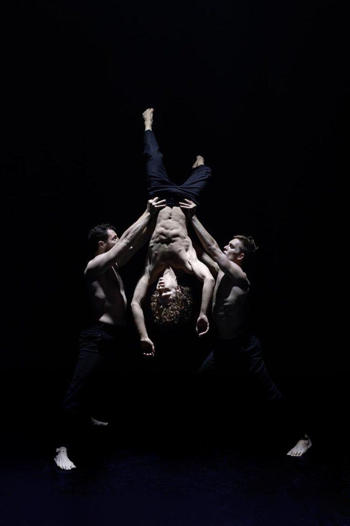 Two male dancers hold another male dancer upside down off the floor by his hips.