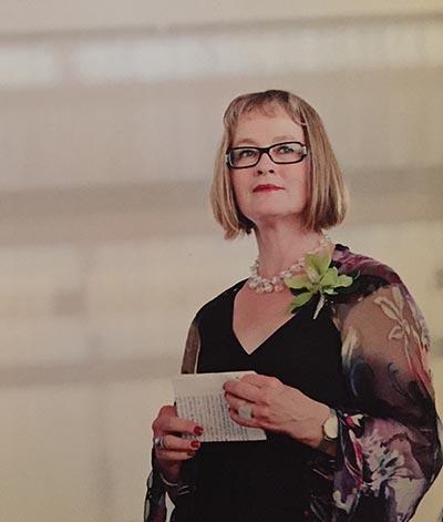 A woman with chin length straight blonde hair with short bangs and glasses stands slightly turned towards the camera. She is wearing a black dress with see-through sleeves with a large flower pattern. She is holding a piece of paper in her hands.