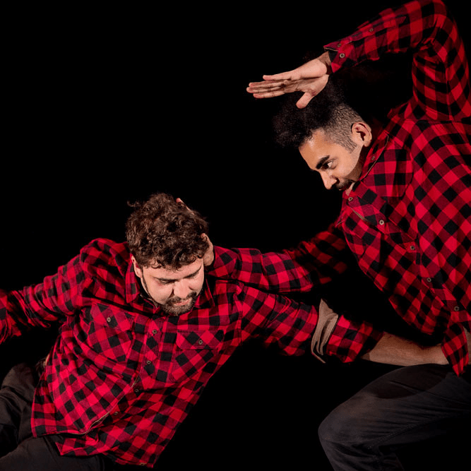 Two men in red and black plaid flannel shirts with dark hair and beards are in frame. One has his left arm raised and bent, with his other hand behind the other man's head. The other man is bent forward with his hand reaching for the first man's hip.