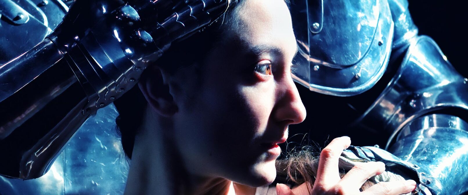 Renée Sigoiun in Kidd Pivot's "Assembly Hall". A close up on Renée's face, looking to the right. A person in a suit of medieval style armour stands close behind with one hand on Renée's head and one on her shoulder.