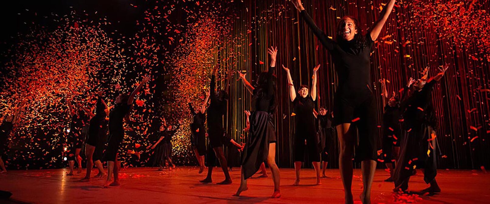 Nova Dance. A scene with a sense of joy that shows a group of dancers spread across a stage all wearing black. Some have their arms raised. The stage is lit red and the backdrop has splashes of red light against black.