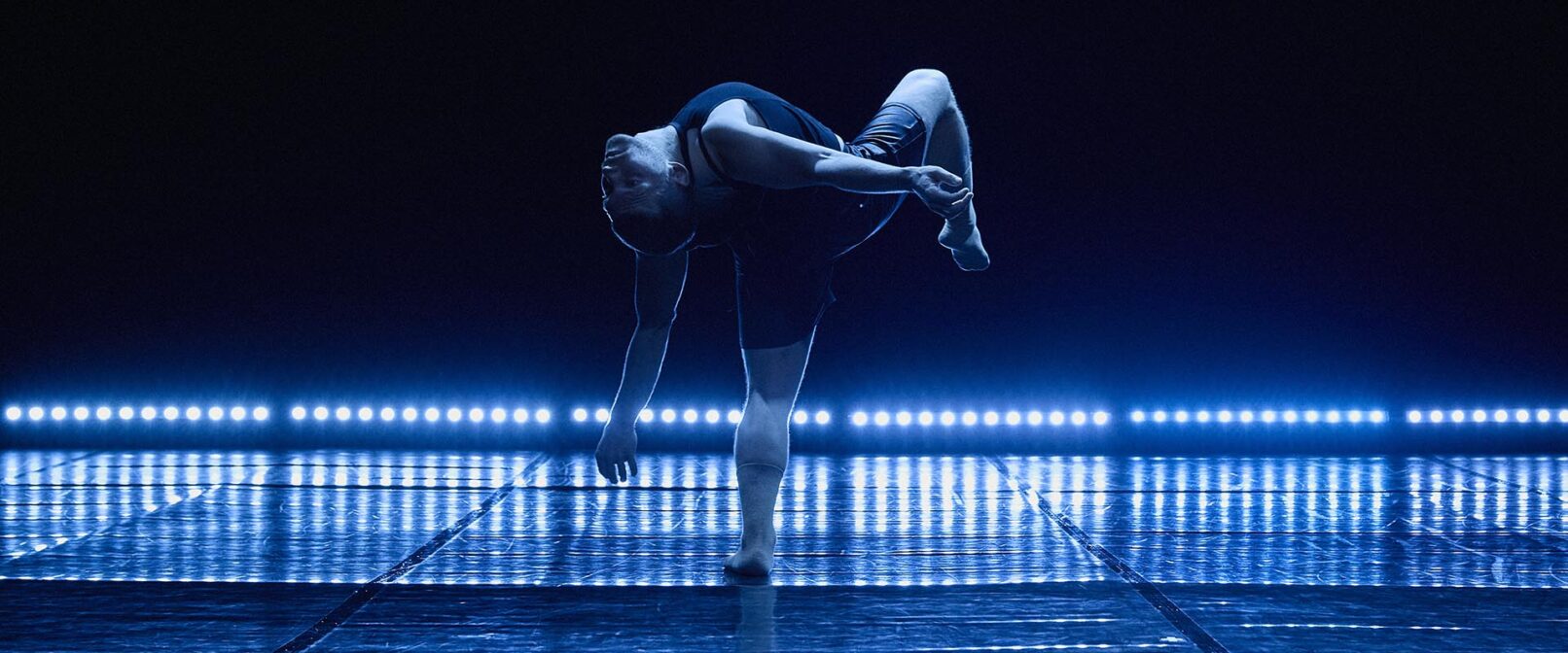 Guillaume Cote Danse. A dancer stands centre stage bending backwards with one leg raised and knee bent. The top half of the background appears dark, while the bottom half is a projection of blue light and shadow forming a row of narrow vertical bars.