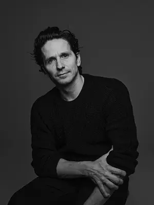 A black and white portrait photo of a light skinned man with short dark hair and a long sleeve black top, bunched up to his elbows. He looks forward with one arm crossed in front of him grabbing his other forearm.