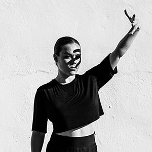 Rocío Molina. A black and white photos of a woman withtightly pulled back black hair stands in front of a white stucco wall. She is wearing a half length sleeve black crop top and black tights. 