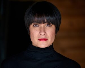 A woman with short dark hair wearing a black turtleneck and red lipstick looking directly at the camera.