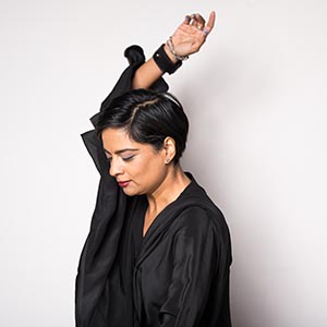 Nova Bhattacharya stands in front of a white wall looking down and turned slightly to her right. Her right arm is raised above her head. She is dressed in a flowing black outfit with black and silver bracelets. Her hair is black and cut short.