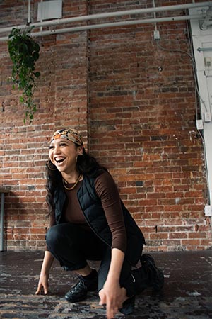 Alyssa Favero. A smiling woman with long, wavy dark hair in black pants, a brown longsleeve top, black puffy vest and a multicolour kerchief on her head crouches in front of a brick wall.