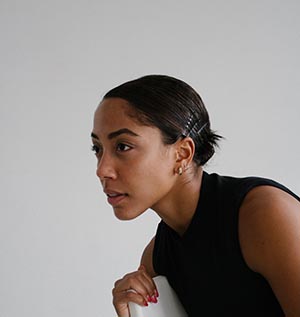 Livona Ellis is pictured sitting, in three quarter profile from the shoulders up, facing to the left. She has short dark hair that is pinned back and is wearing a sleeveless black top.