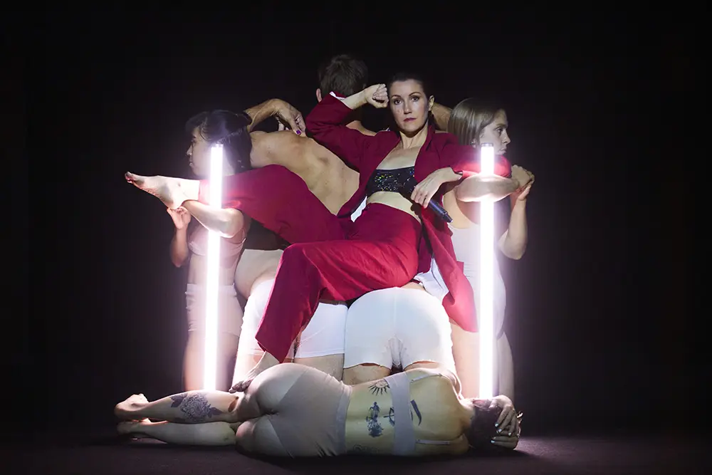 A woman in red pants and a red blazer opened down the front to reveal a patterned sports bra sits on a group of dancers who are kneeling to form a chair-like structure with their bodies. A female dancer with tattoos in beige coloured bra and panties lies on the ground with her back to the camera.