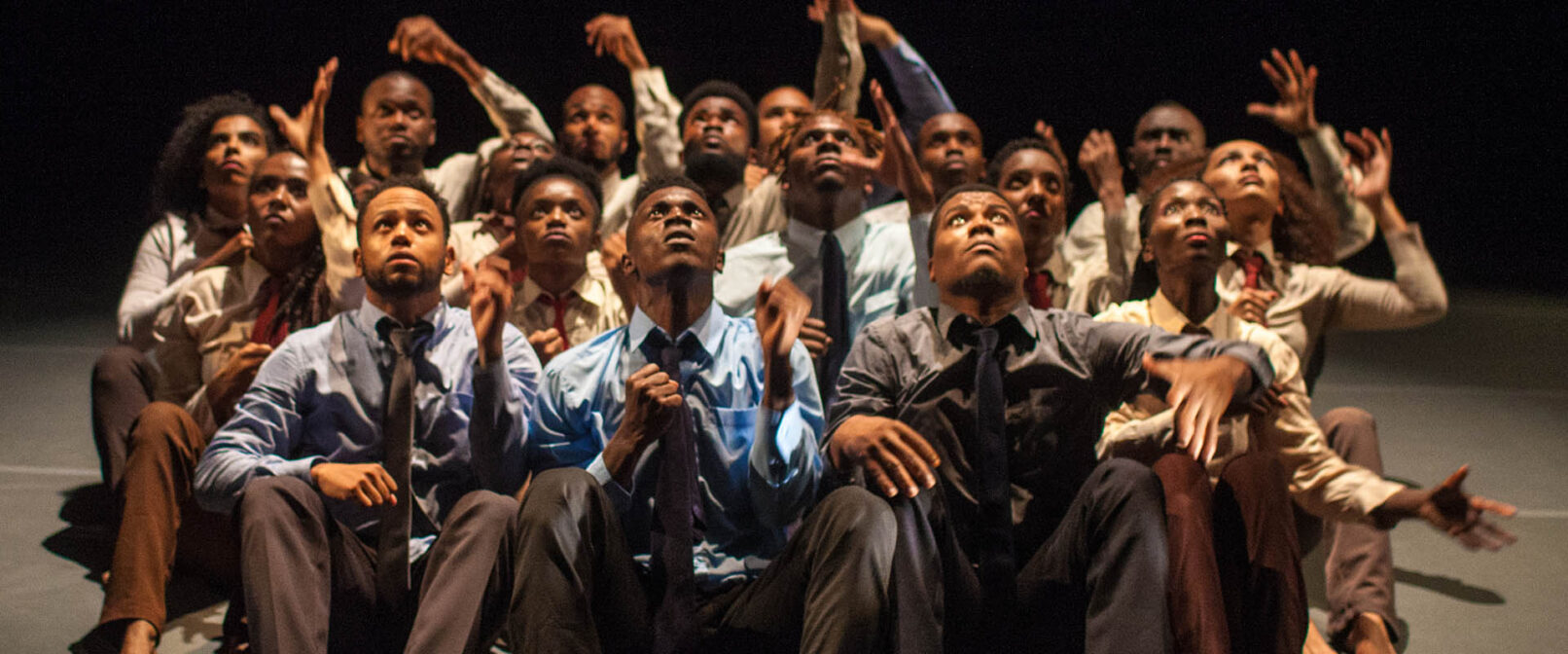 Sankofa Danzafro. A group of seventeen Black dancers of mixed genders sit together on the floor facing forward, looking up in the air. Some have arms reaching upwards. They are all dressed in button up tops with ties and slacks.