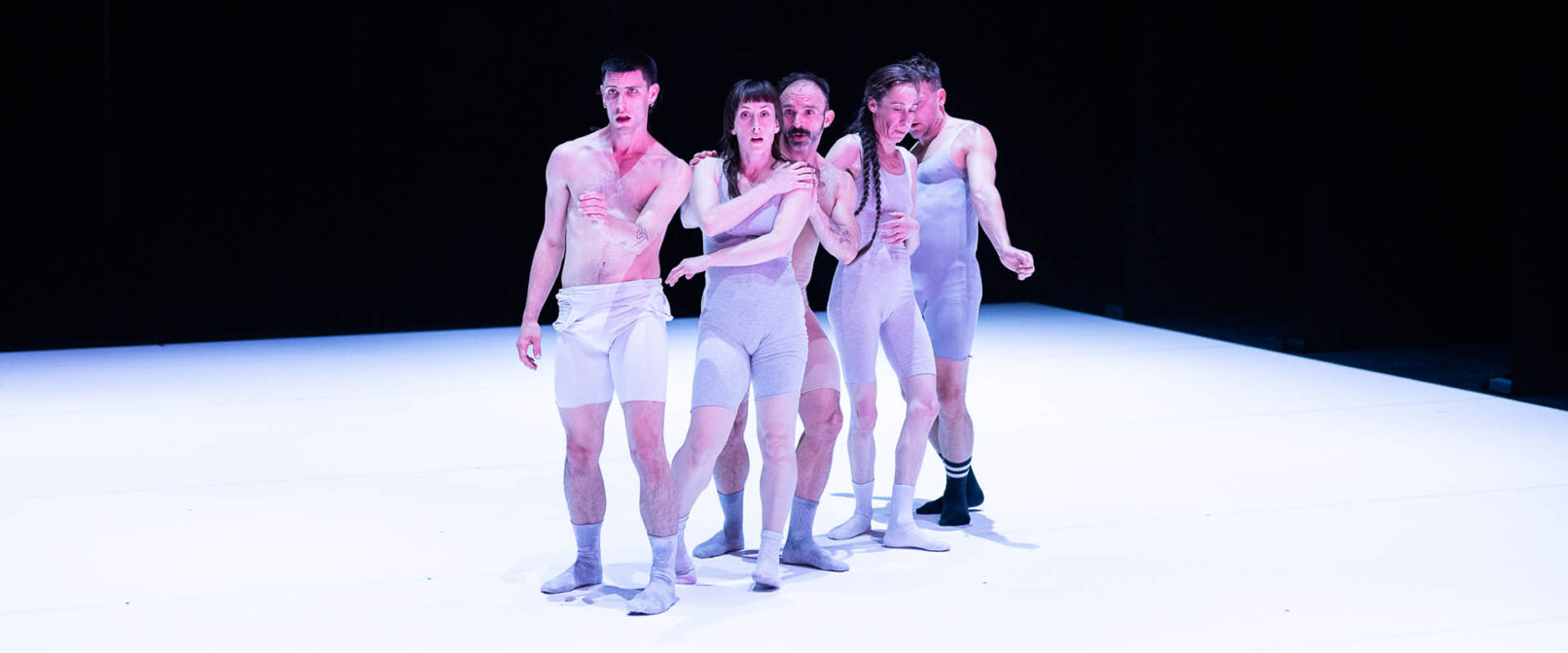 Compagnie Catherine Gaudet. Five fair skinned dancers stand grouped tightly together on a white stage, facing forward. They are wearing white and light grey tight sleeveless, and thigh length body suits.