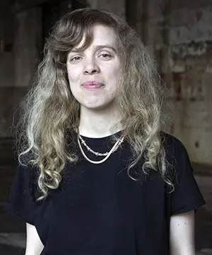 A fair skinned woman with long, wavy blond and light brown hair smiles facing forward. She is wearing a black t-shirt and beaded necklaces.