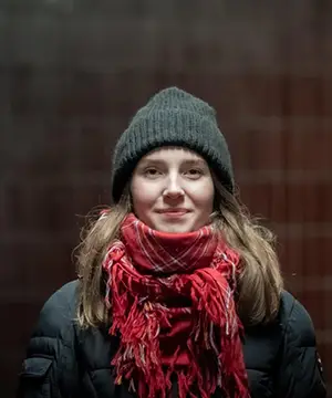 A fair skinned woman with light brown hair faces the camera. She is wearing a grey toque and red plaid scarf.