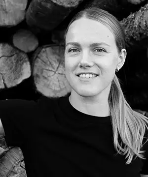 A black and white photo of a fair skinned woman with long dirty blond hair tied in a low ponytail. She faces the camera smiling. She is wearing a black top and is in front of a pile of cut timber ends.