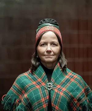 A fair skinned woman with shoulder length brown hair faces forward. She is wearing a traditional Samí hat in green and red and a green adn red plaid shawl with a circle clasp at the chest.