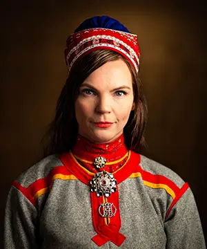 A fair skinned woman with long brown hair faces the camera. She is wearing a traditional Samí hat and tunic in grey with red and yellow strips at the shoulders and down the chest.