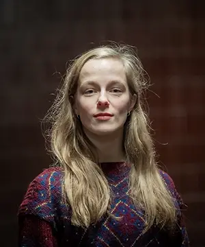 A fair skinned woman with long blond hair faces forward. She is wearing a red and blue patterned sweater.