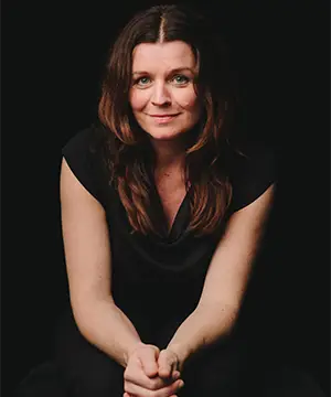 A fair skinned woman with long brown hair and a black t-shirt sits facing forward with her hands clasped on her knees.