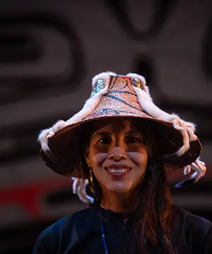 A portrait photo of Margaret Grenier, A woman with long brown hair wearing a woven cedar bark hat and black shirt.